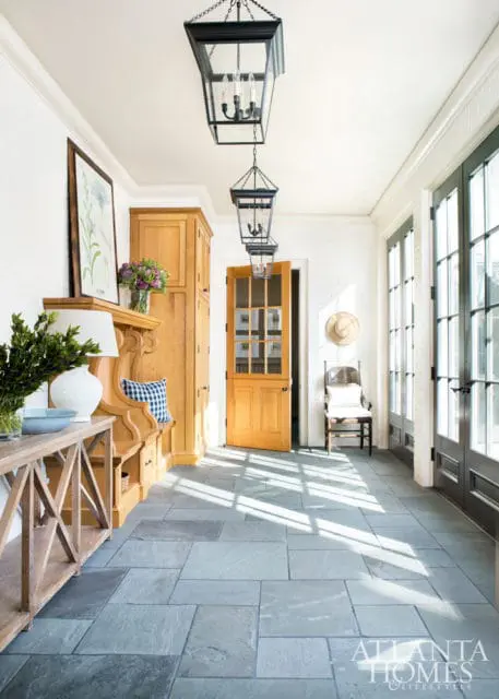 Sunlight streams across a hallway with blue slate tile floors, white brick walls, and a wooden built-in bench with storage. Black lantern pendant lights hang from the ceiling.
