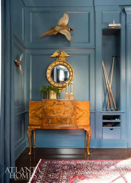 Blue paneled walls with a taxidermied pheasant, a gold eagle mirror, a wooden cabinet, and a Persian rug.