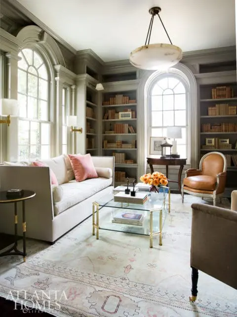 Library with floor-to-ceiling bookshelves, arched windows, a cream sofa with pink pillows, and a glass and brass coffee table.
