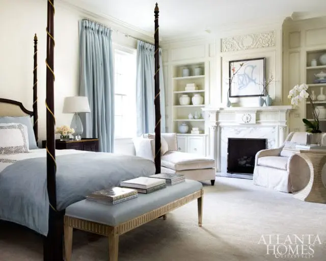 Bedroom with a dark wood four-poster bed, light blue bedding, and a pale blue bench with gold accents. A fireplace with marble surround and ornate mantelpiece is visible, flanked by bookshelves.