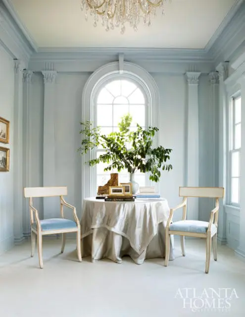 Light blue room with a round table draped in a champagne-colored cloth, two cream-colored chairs with light blue velvet seats, and an arched window with a crystal chandelier above.