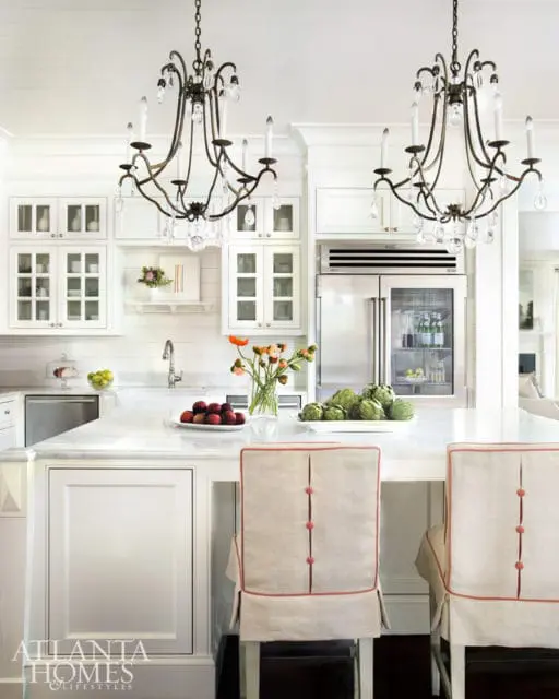 White kitchen with marble island, two ornate chandeliers, stainless steel refrigerator, and upholstered chairs with coral trim.
