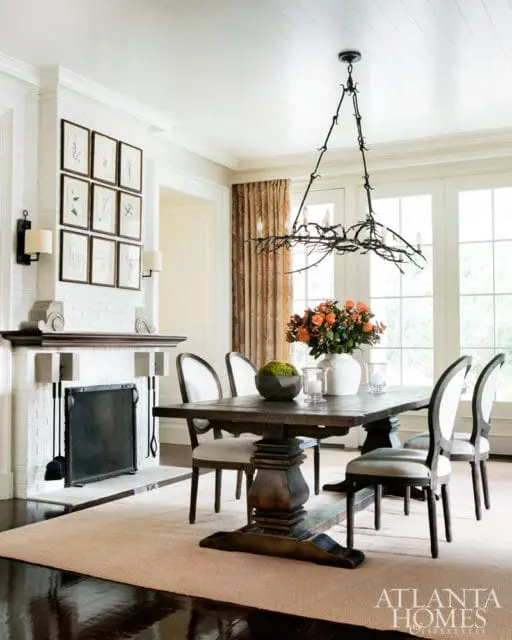 Dining room with a dark wood trestle table and upholstered chairs, white brick fireplace with artwork, and a branch-like chandelier.