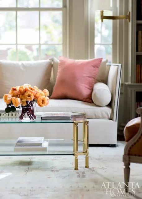 Living room with a cream sofa, coral throw pillow, glass coffee table with books and flowers, and a patterned rug.