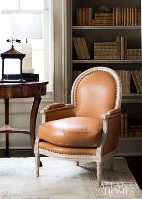 Brown leather armchair with nailhead trim in front of a bookshelf filled with vintage books. A round wooden side table with a lamp sits to the left.