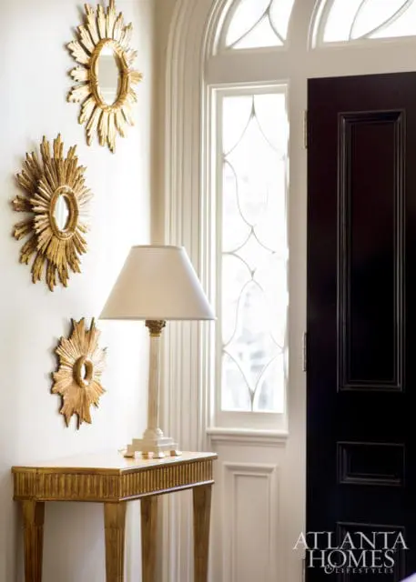 Entryway with three gold sunburst mirrors on a white wall, a gold fluted console table with a cream lamp, and a dark wood door next to a tall arched window.