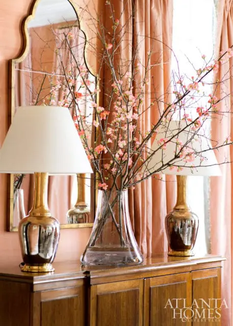 Wooden sideboard with two brass lamps, a glass vase of flowering branches, and a scalloped mirror reflecting peach curtains.