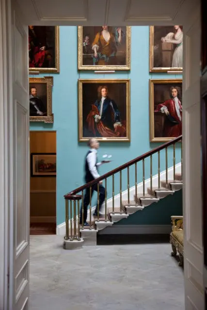 Man in a suit carrying a tray up a carpeted staircase with ornate railings, flanked by classical portraits on a teal wall.