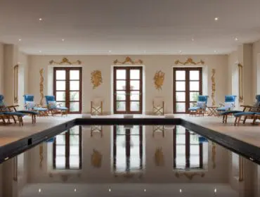 Indoor swimming pool with dark water, surrounded by light-colored floor and walls, featuring multiple wooden lounge chairs with blue cushions and decorative gold wall accents.