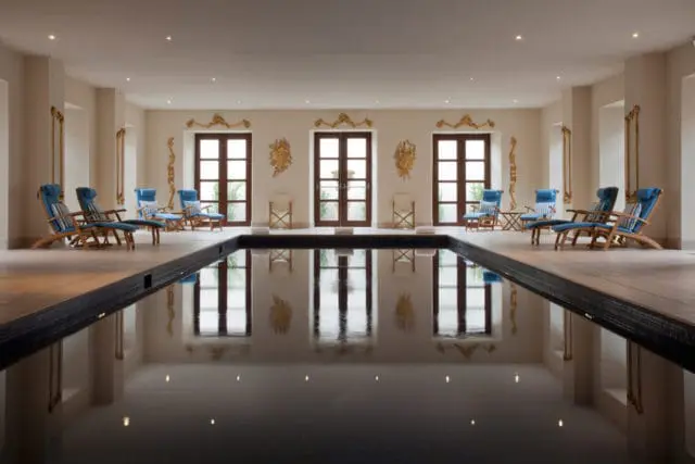 Indoor swimming pool with dark water, surrounded by beige walls, wooden doors, and blue lounge chairs with striped cushions.