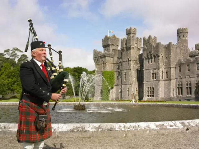 Man in kilt and suit playing bagpipes in front of a stone castle with a fountain and pond