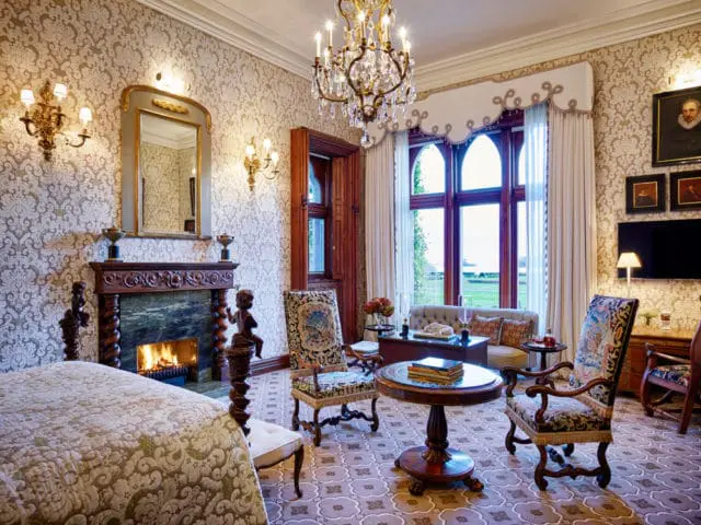 Ornate bedroom with a fireplace, patterned wallpaper, crystal chandelier, and antique furniture. A large window overlooks a green landscape.