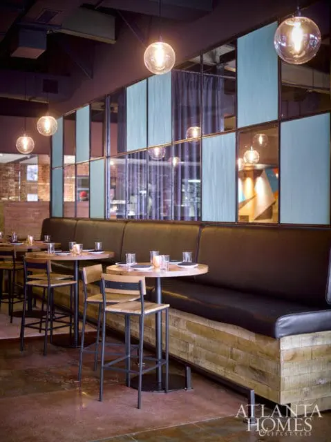 Restaurant dining area with dark leather banquette seating, round wooden tables, and industrial-style pendant lights. A decorative glass partition with blue panels separates the seating area.