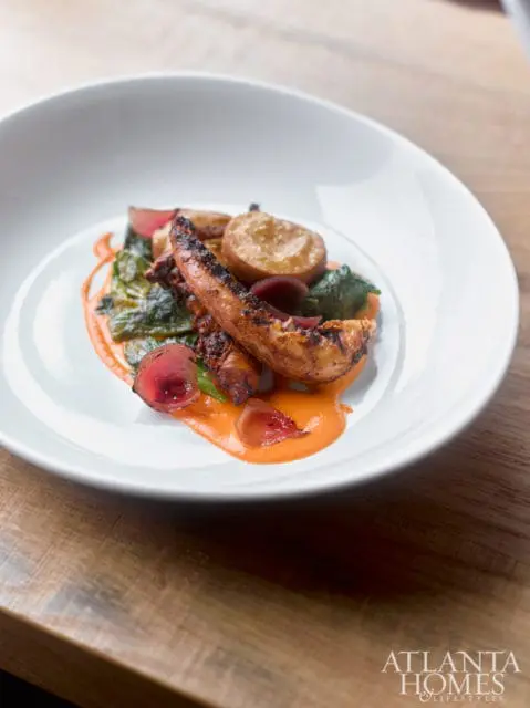 Grilled octopus tentacles served with roasted potatoes, greens, and a vibrant orange sauce on a white plate.