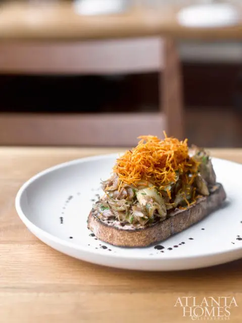 Toasted sourdough bread topped with shredded vegetables and chopped herbs, served on a white plate with a drizzle of dark sauce.