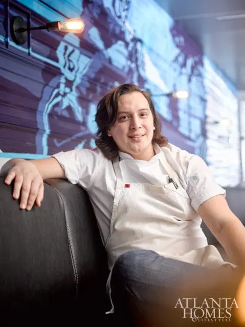 Chef in white chef coat and apron sitting on a dark leather couch in front of a blue mural and industrial lights.