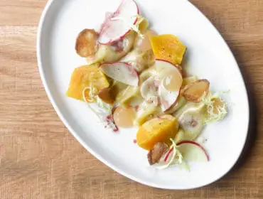 White oval plate with pasta, roasted butternut squash, sliced apples, radishes, and crispy potato chips on a wooden table
