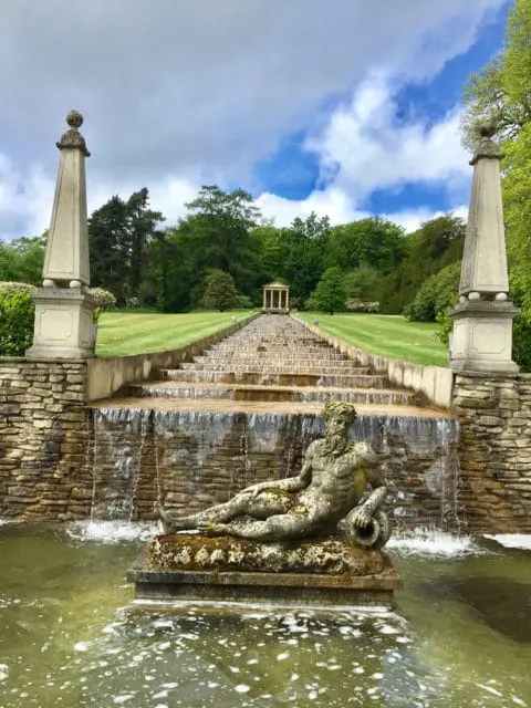 Stone statue of a reclining man in a pond at the base of a tiered waterfall and staircase, flanked by obelisks and leading to a classical temple structure.