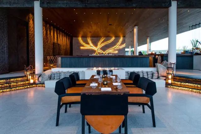 Dining room with a long wooden table set for a meal, featuring black and orange chairs, a central fireplace, and a large illuminated branch art piece on the back wall.