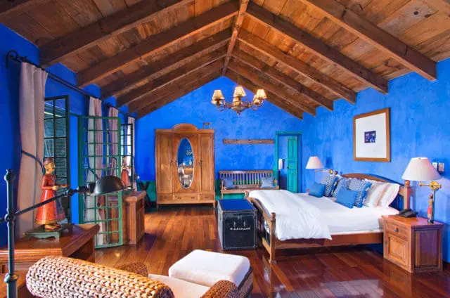 Bedroom with vibrant blue walls, vaulted wood-beamed ceiling, and polished wood floors. Features a wooden bed with blue pillows, an ornate armoire, and decorative figures.