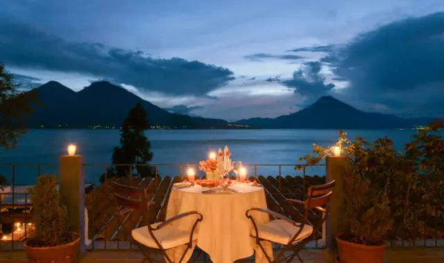 Table set for two with candles and wine glasses on a balcony overlooking a lake and mountains at dusk