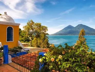 Yellow building with white dome overlooks a bright blue lake with two volcanic mountains in the distance under a clear sky.