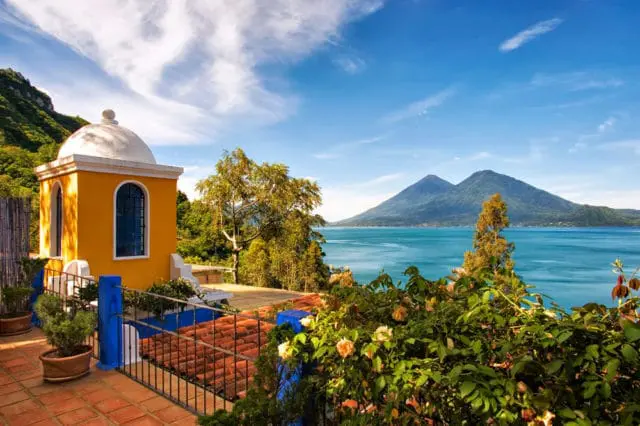 Terracotta tiled patio with a yellow domed structure overlooks a bright blue lake with two volcanic mountains in the distance under a cloudy sky.