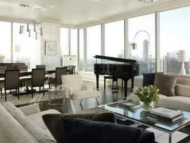 Luxury living and dining room with a grand piano, large dining table, and sectional sofas, featuring floor-to-ceiling windows with city views.