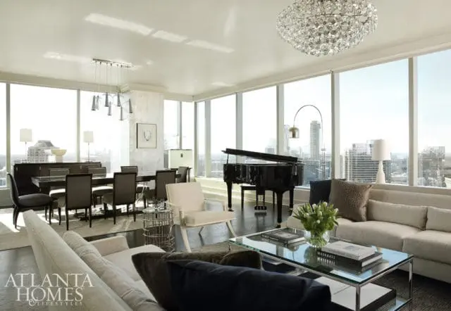Spacious living and dining room with a grand piano, large windows overlooking a city skyline, a crystal chandelier, and modern furniture.