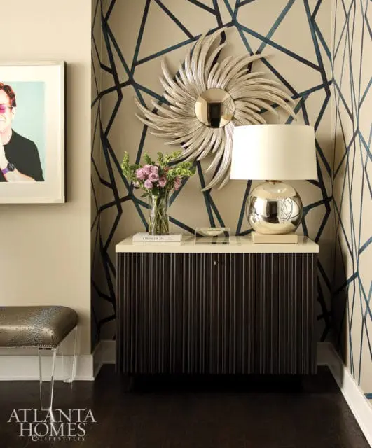 Hallway with geometric wallpaper, a sunburst mirror, a silver lamp on a dark fluted cabinet, and a framed portrait of Elton John.
