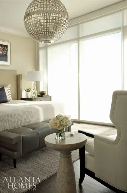 Bedroom with a large orb chandelier, upholstered headboard, bedside table with lamp, gray leather bench, and a small pedestal table with a vase of white roses.