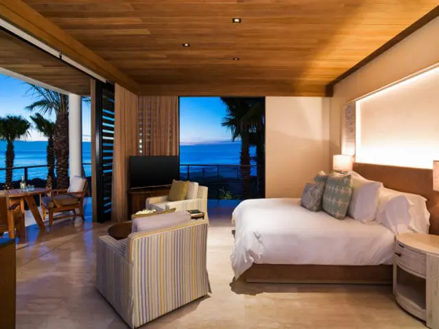 Bedroom with a king-size bed, upholstered chairs, and a television, opening to a balcony with ocean views at dusk.