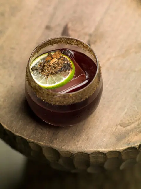 Close-up of a dark red cocktail in a glass with a salted rim, garnished with a lime wheel dusted with spices and a large ice cube.