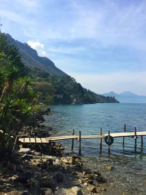 Wooden dock extending into clear blue water with rocky shore and lush green mountainside under a bright blue sky with scattered clouds.