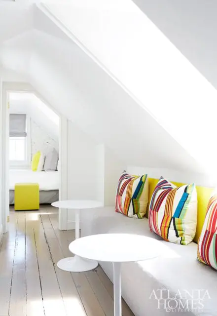 Attic room with a built-in sofa featuring colorful striped pillows and a yellow backrest, two white tulip tables, and a doorway leading to a bedroom with a yellow ottoman.