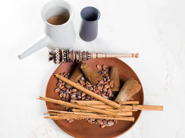 Clay plate with cacao beans, cinnamon sticks, and piloncillo sugar cones, next to a molinillo whisk and a jug of hot chocolate.