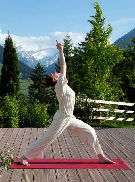 Woman in beige yoga attire performs Virabhadrasana II pose on a red mat on a wooden deck, with a backdrop of green trees and snow-capped mountains.