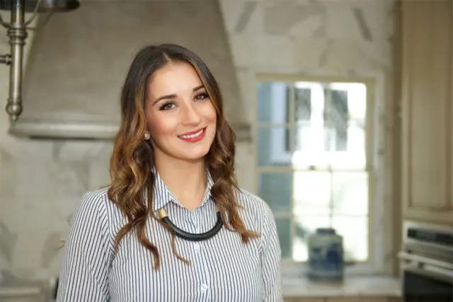 Woman with wavy brown hair wearing a striped shirt and a black necklace smiles in a kitchen with marble countertops and a window in the background.