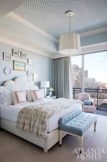 Light blue bedroom with a white cloud-shaped headboard, white bedding with plush throw, blue tufted bench, patterned curtains, and a blue and white checkered ceiling.
