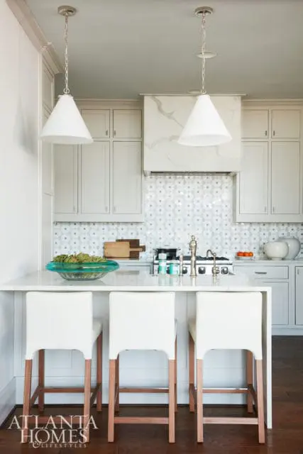 Light gray kitchen with white marble waterfall island, white bar stools with wood legs, and geometric tile backsplash.