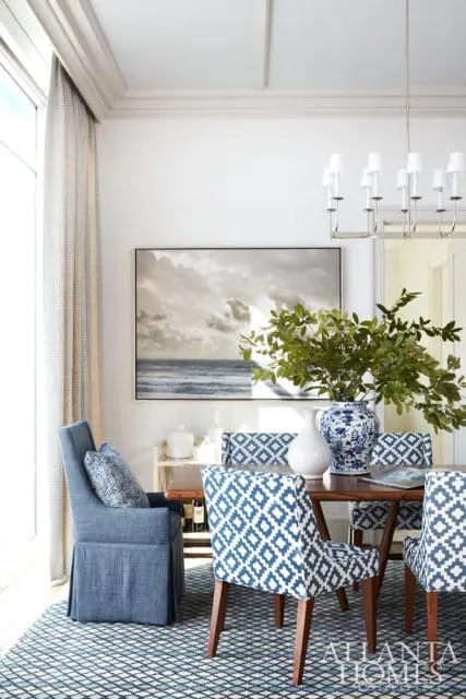 Dining room with blue and white patterned chairs, a blue slipcovered armchair, a dark wood table, and a large framed seascape artwork above a console table.