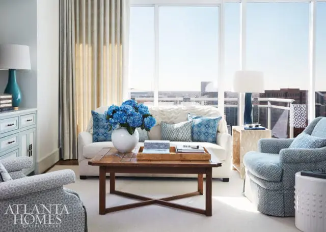Living room with white sofa and chairs upholstered in blue and white patterned fabric, a wooden coffee table, and large windows overlooking a city skyline.