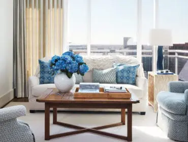 Living room with white sofa, blue patterned accent pillows, and a wooden coffee table with a vase of blue hydrangeas.