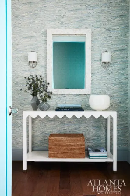 Entryway with blue wave-patterned wallpaper, a white scalloped console table, a woven basket, and a mirror with a mother-of-pearl frame.