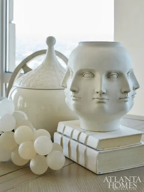 White multi-faced bust vase on stacked white books, next to a large white lidded jar and a bunch of white grapes.