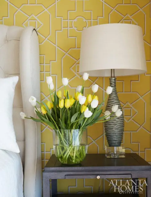 Bedroom with yellow geometric wallpaper, tufted headboard, and a bedside table with a glass vase of yellow and white tulips and a textured lamp.