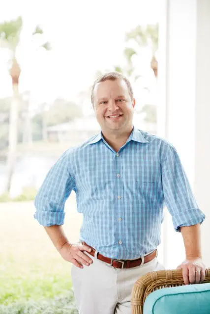 Man in blue plaid shirt and white pants with hands on hips, leaning on wicker chair with teal cushion. Palm trees and water visible in background.