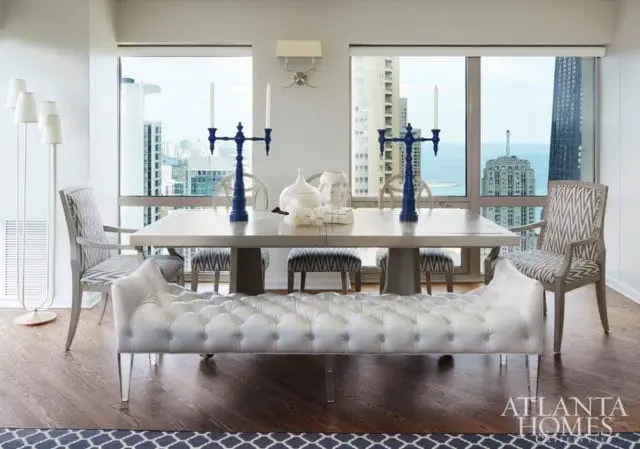 Dining room with a light wood table, white tufted bench, upholstered chairs with geometric pattern, and large windows overlooking a city skyline.