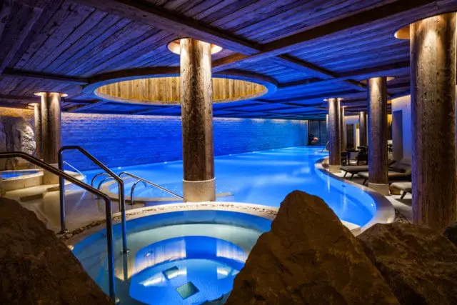 Indoor spa pool with bright blue water, surrounded by stone walls, wooden beams on the ceiling, and large natural rock features.