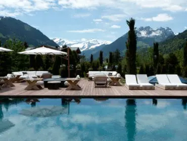 Outdoor swimming pool with lounge chairs and seating area on a wooden deck, with snow-capped mountains and trees in the background.